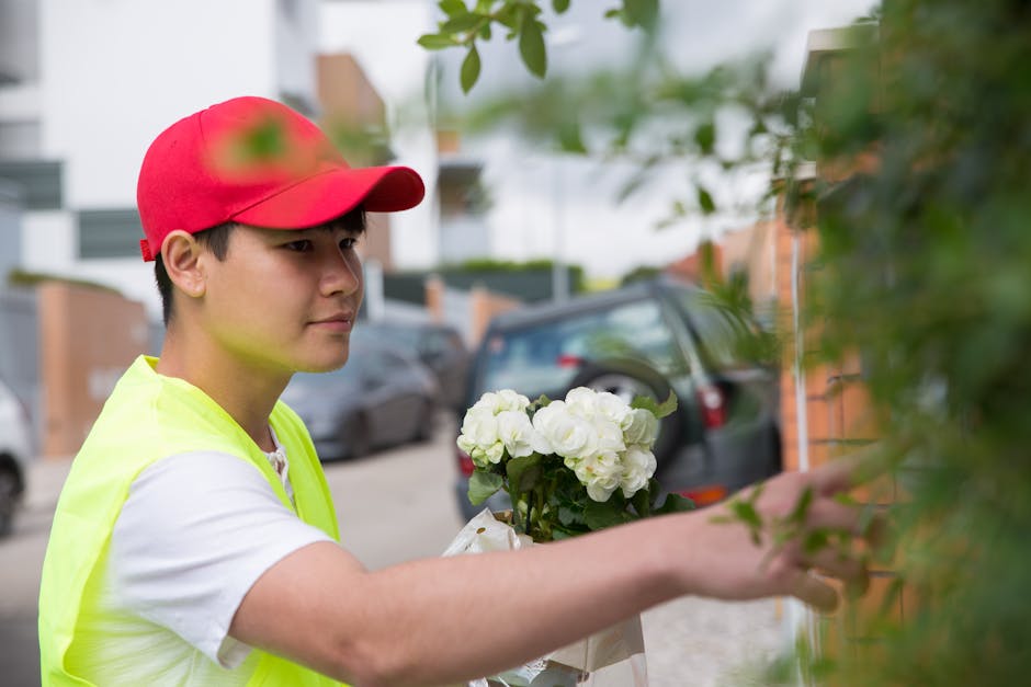A young male florist wearing a red cap and a high-visibility yellow vest arranges a bouquet of white hydrangeas, carefully placing the flowers against a shrub on a residential street. The bouquet is w