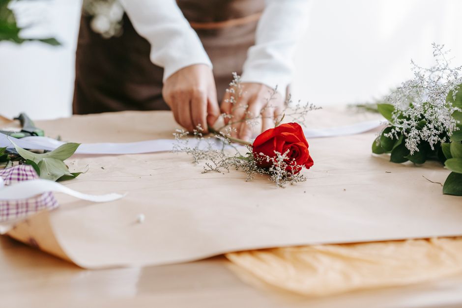 A person arranging a floral bouquet on a wooden table, featuring a vibrant red rose at the center, surrounded by delicate white filler flowers and lush green foliage. The individual is wearing a white