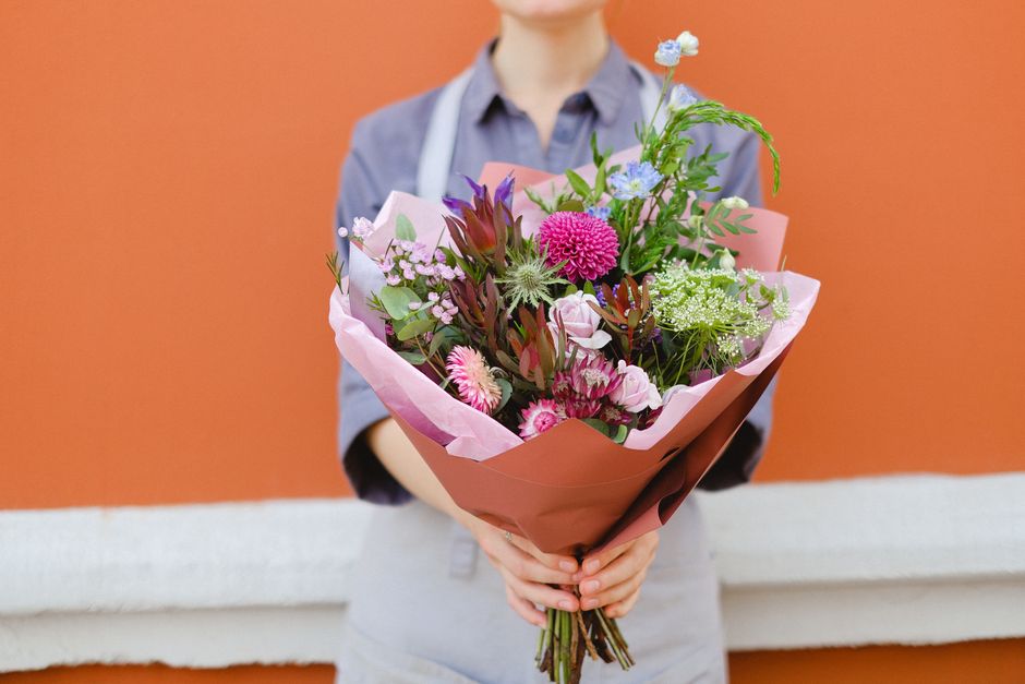 A person dressed in a light grey shirt and apron, holding a vibrant floral bouquet in front of them. The bouquet features a mix of fresh flowers including pink and purple dahlias, blue cornflowers, pi