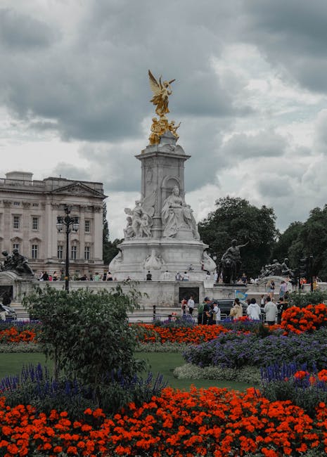 A view of the Victoria Memorial with its ornate marble sculpture and gilded angel atop, surrounded by well-maintained flower beds featuring vibrant red, purple, and green foliage. The flowers include 