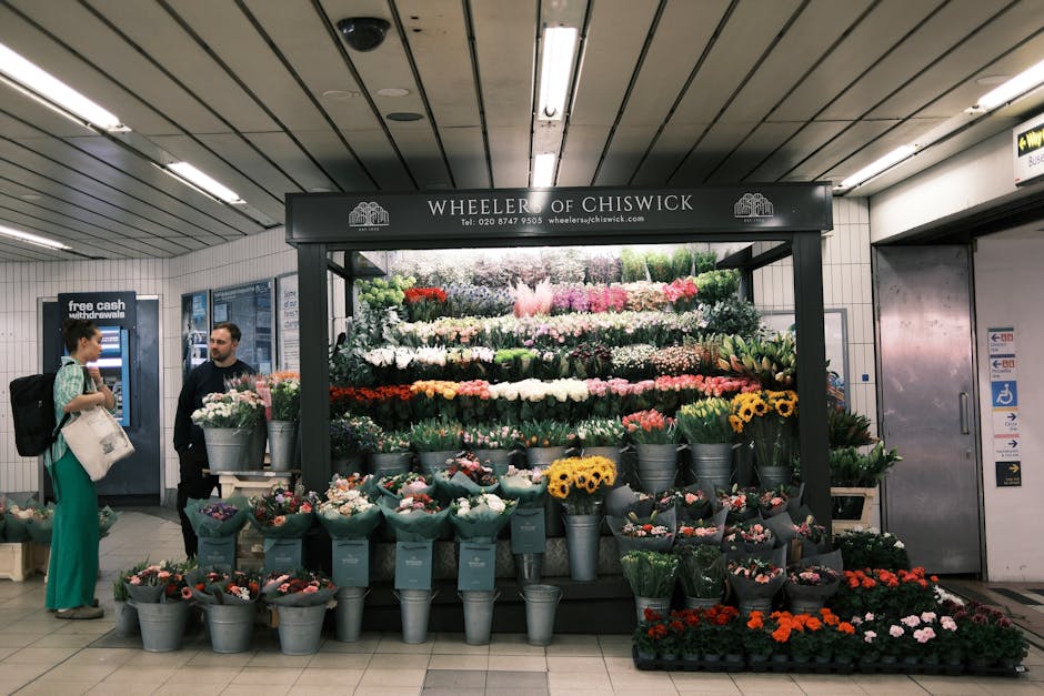 Inside an underground station, a flower stall named Wheeler of Chiswick displays a vibrant floral arrangement. The stall is filled with a variety of fresh flowers, including pink, white, red, yellow, 