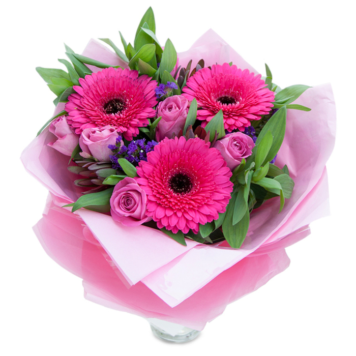 I see this bouquet as a little celebration of pinks and purples, composed with the craft of a neighbourhood florist who knows Chelsea's love of refined colour. At the heart are three striking fuchsia Gerbera daisies, their layered petals splayed like miniature sunbursts and centred by dark discs that provide a confident visual punctuation. Surrounding them, roses in a dusty lavender-pink curl inward in soft, velvety spirals; their budlike shape suggests freshness and offers a gentle, rose-sweet aroma that mingles subtly with the green notes of foliage. Intermittent sprays of purple statice drift through the composition, lending a delicate, papery texture and a cool violet thread that balances the warmer pinks. The supporting greenery-broad salal or eucalyptus-like leaves-creates a deep verdant chorus, offering both structure and a crisp, slightly resinous scent that evokes morning walks past local Chelsea gardens. The bouquet is wrapped artfully: an inner sheet of matte pale pink tissue cushions the stems, while an outer layer of translucent, slightly crinkled fuchsia-pink cellophane catches the light and ties the colour story together. The lower portion reveals the top rim of a clear glass vase, indicating the arrangement is ready to grace a mantel or kitchen counter. Soft, even lighting brings out the saturations of pink and the subtle sheen on each petal. As prepared for delivery within Chelsea-perhaps to a flat near the King's Road or tucked into a bedside table on Flood Street-this bouquet feels intimate, celebratory, and quietly luxurious.